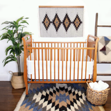 Wooden crib in a cozy nursery with a bold Smilyv Wool Rug / Wall Hanging in neutral hues, a potted plant, basket, and geometric wall art.