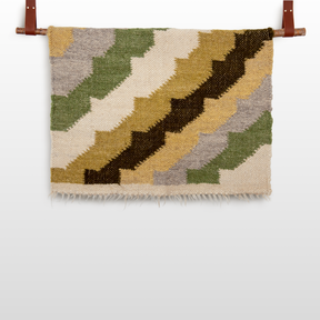 The Sontse (Sun) Wool Statement Throw / Baby Blanket is a handcrafted wool blanket with zigzag patterns in green, beige, brown, and cream, displayed hanging from two leather straps.