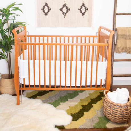 The Sontse (Sun) Wool Statement Throw Baby Blanket is draped over a wooden crib with white bedding, set on a patterned rug next to a plant and a basket with towels and a linen laundry bag.