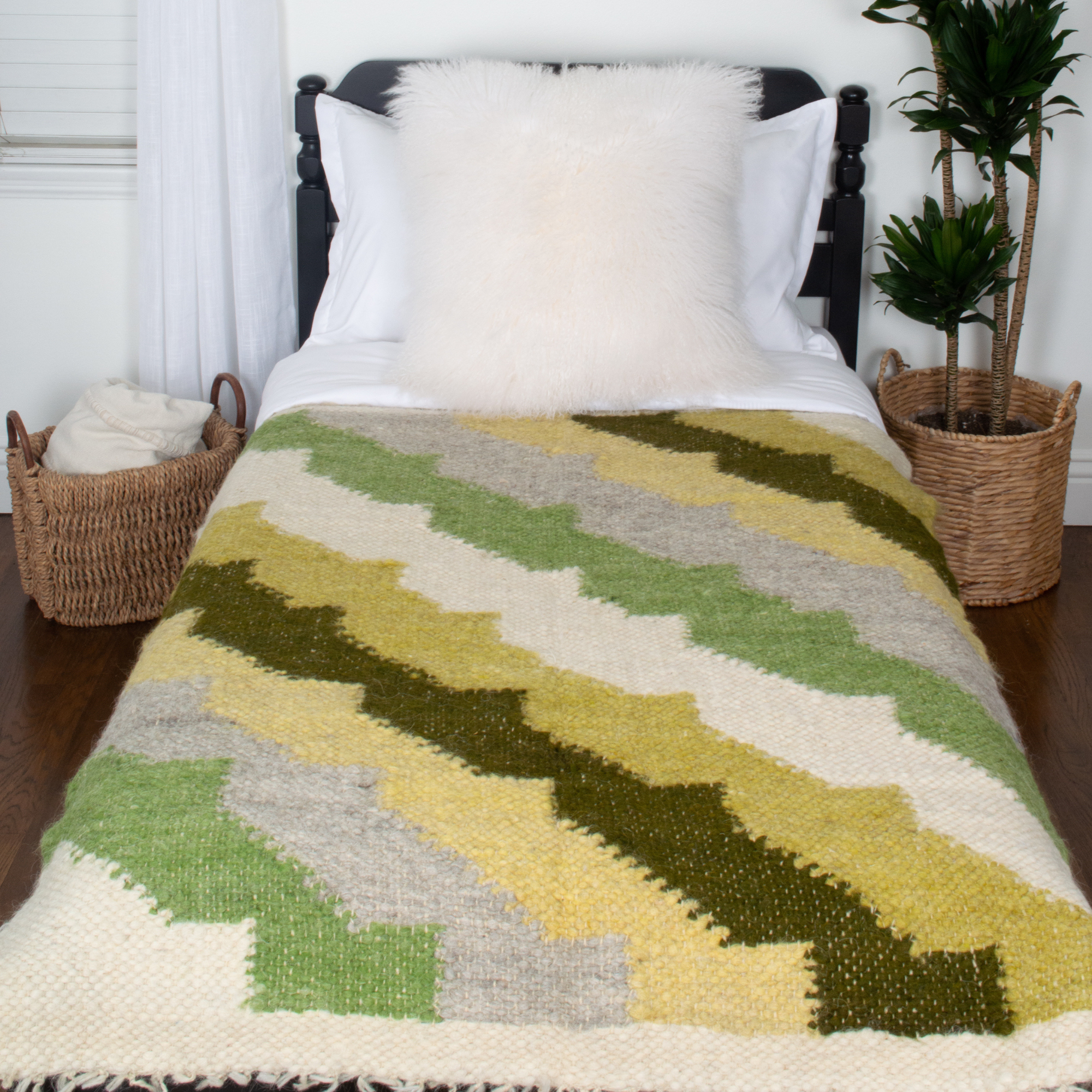 A single bed features the Sontse (Sun) Wool Statement Throw / Baby Blanket in green and beige zigzags, a white pillow, and baskets beside it on a wooden floor.