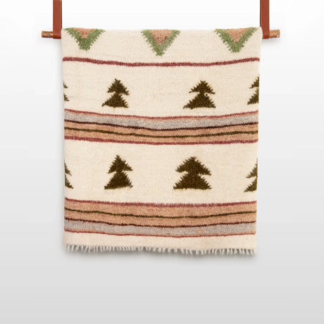 The Sosna (Pine) Wool Statement Bed Blanket is a beige, organic wool blanket with green, brown, and rust tree and stripe patterns, shown hanging on a wooden rod.