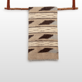 Tak (Yes) Wool Statement Bed Blanket in beige and brown pattern displayed draped over a wooden rod with leather straps, set against a white background.