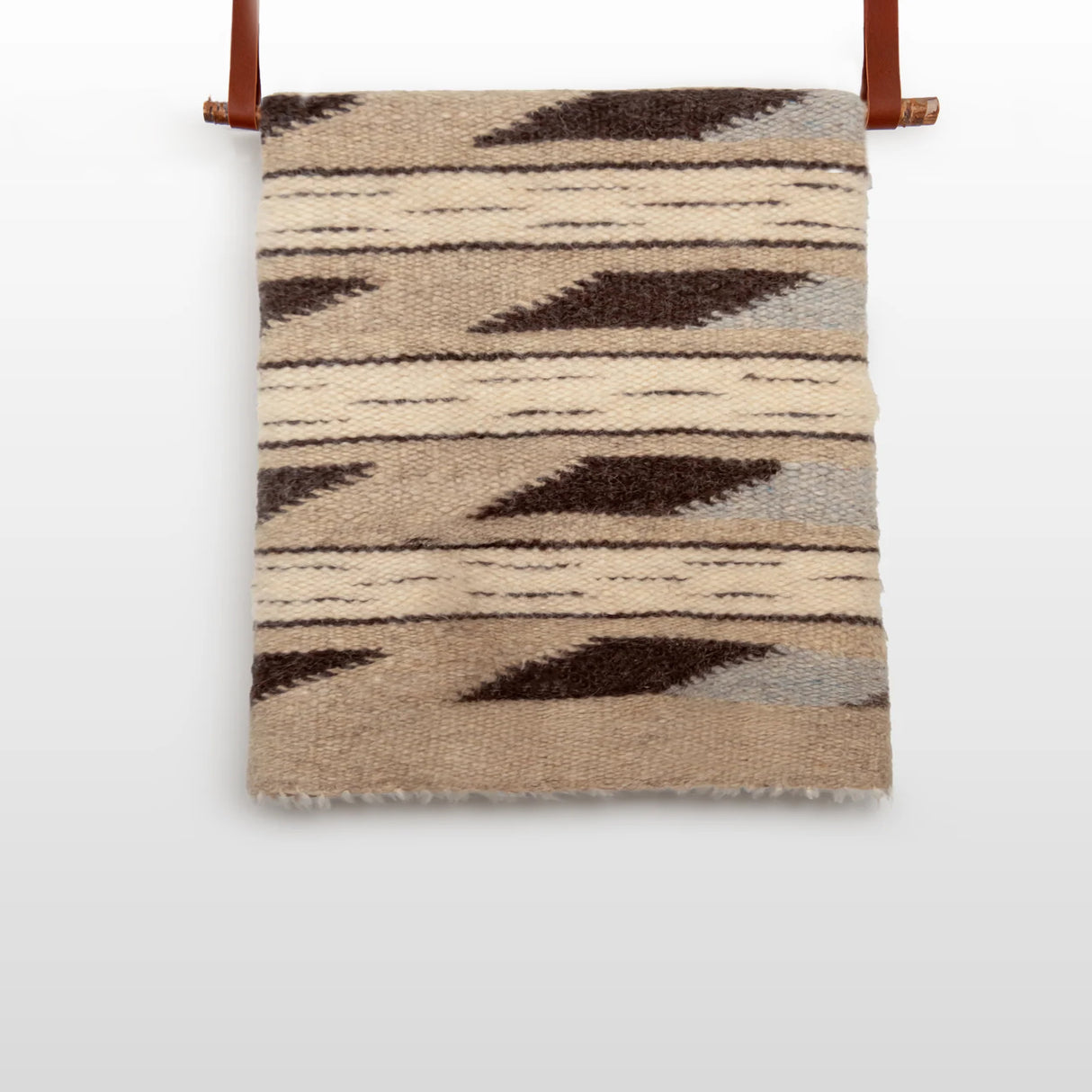 The Tak (Yes) Wool Statement Bed Blanket features beige and brown geometric patterns, handwoven from Carpathian sheep wool, and is displayed on a wooden rod with leather straps.