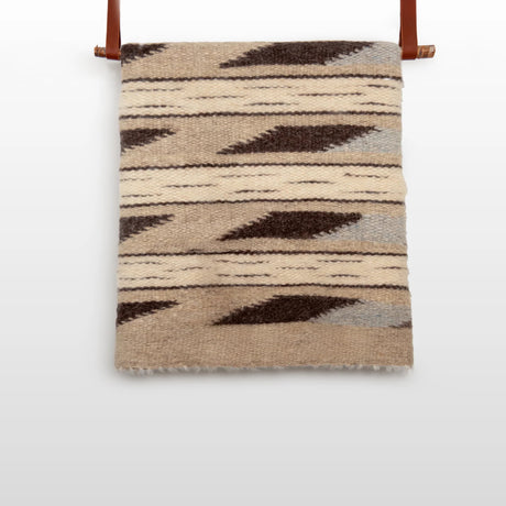 The Tak (Yes) Wool Statement Bed Blanket features beige and brown geometric patterns, handwoven from Carpathian sheep wool, and is displayed on a wooden rod with leather straps.