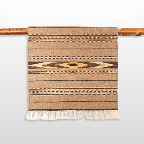 The Teplota (Warmth) Wool Small Rug / Mat - Dark features brown geometric patterns and fringe, displayed on a wooden rod—ideal as a distinctive piece of wall decor.
