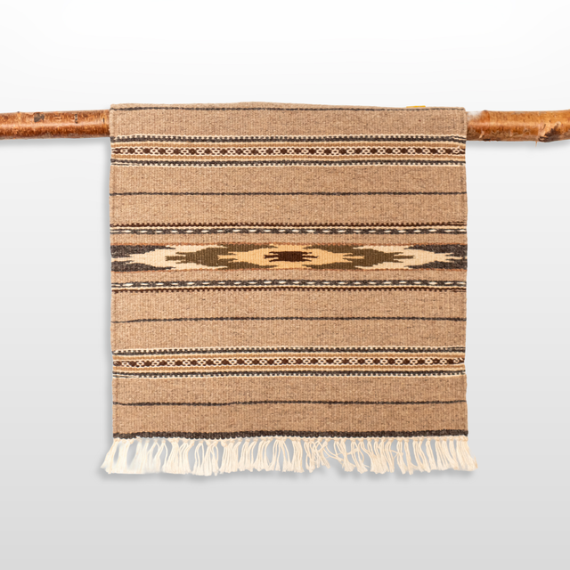 The Teplota (Warmth) Wool Small Rug / Mat - Dark features brown geometric patterns and fringe, displayed on a wooden rod—ideal as a distinctive piece of wall decor.