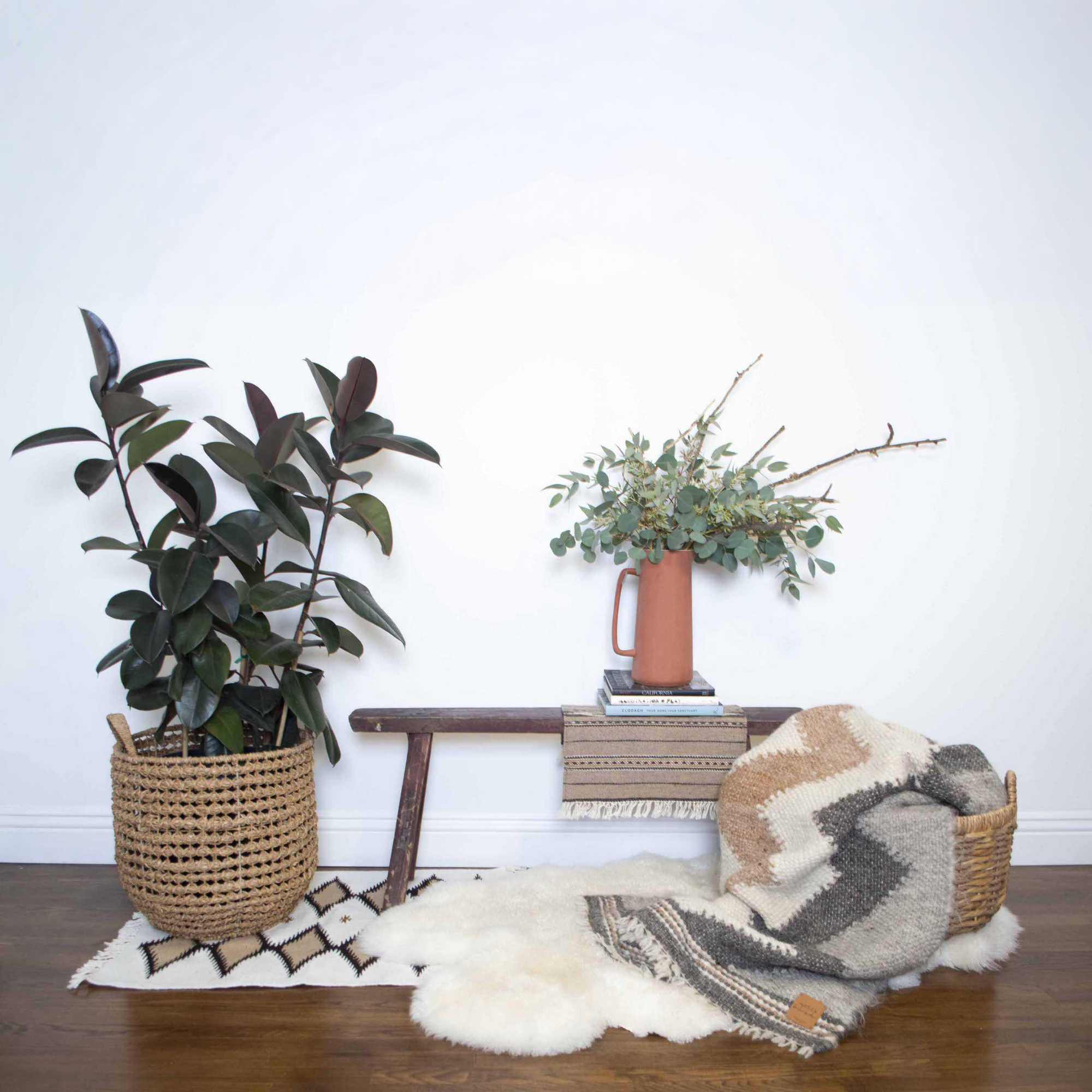 Boho decor with a potted plant, wooden bench, books, wall art, vase of greenery, a cozy blanket, and the Teplota (Warmth) Wool Small Rug / Mat - Dark.