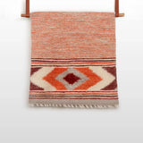 The Tkach (Weaver) Wool Statement Bed Blanket, featuring red, orange, and cream geometric patterns, hangs on a rod with leather straps.