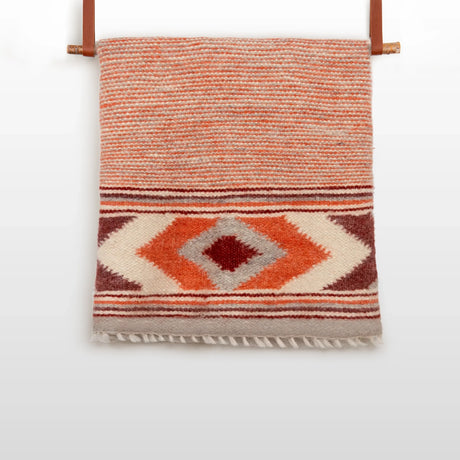 The Tkach (Weaver) Wool Statement Bed Blanket, featuring red, orange, and cream geometric patterns, hangs on a rod with leather straps.