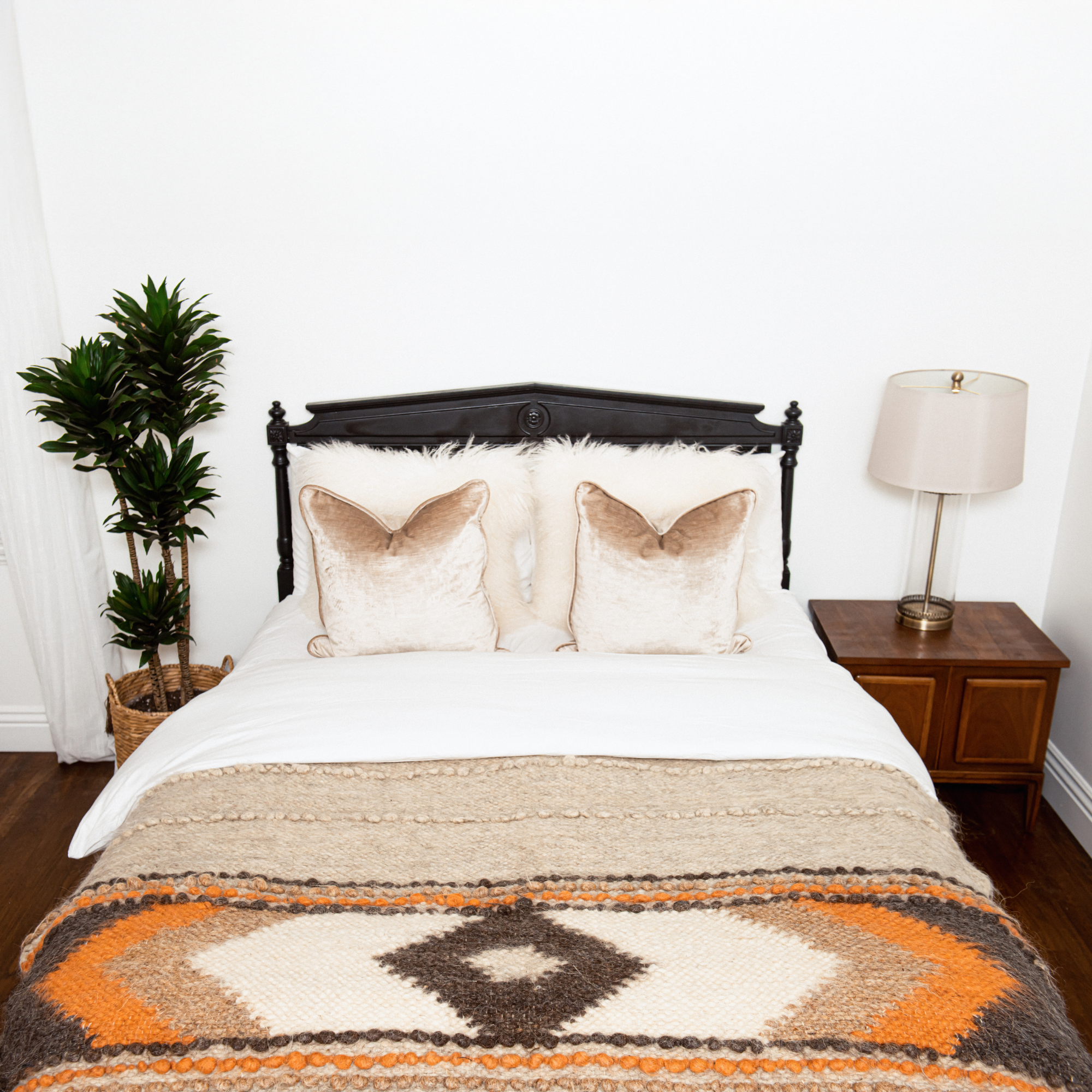 A neatly made bed with beige and orange bedding, two pillows, a lamp, a plant, and a wooden nightstand, topped with the Tkach (Weaver) Wool Statement Bed Blanket for a cozy touch.