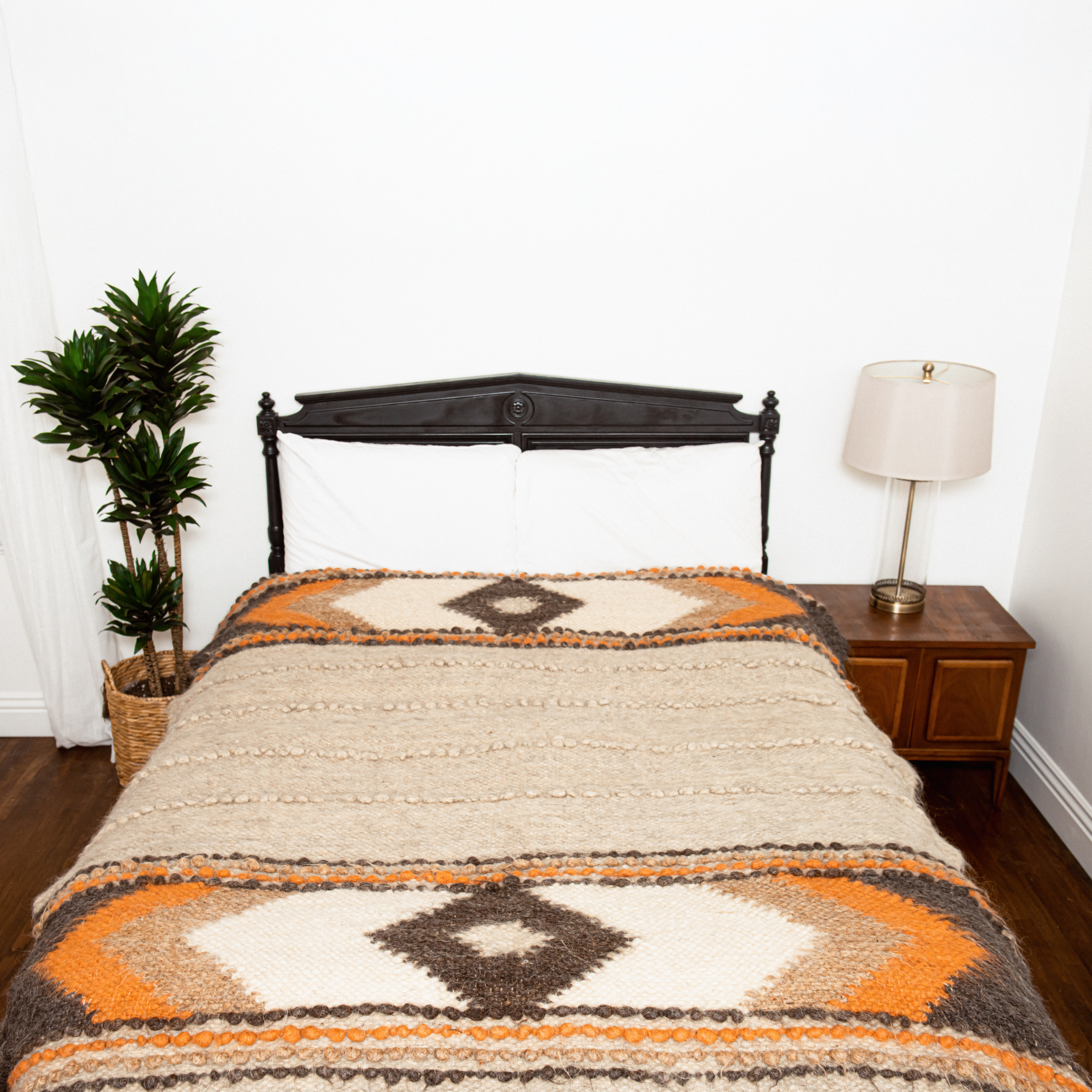A bed with the Tkach (Weaver) Wool Statement Bed Blanket, a nightstand with a lamp, and a potted plant beside it.