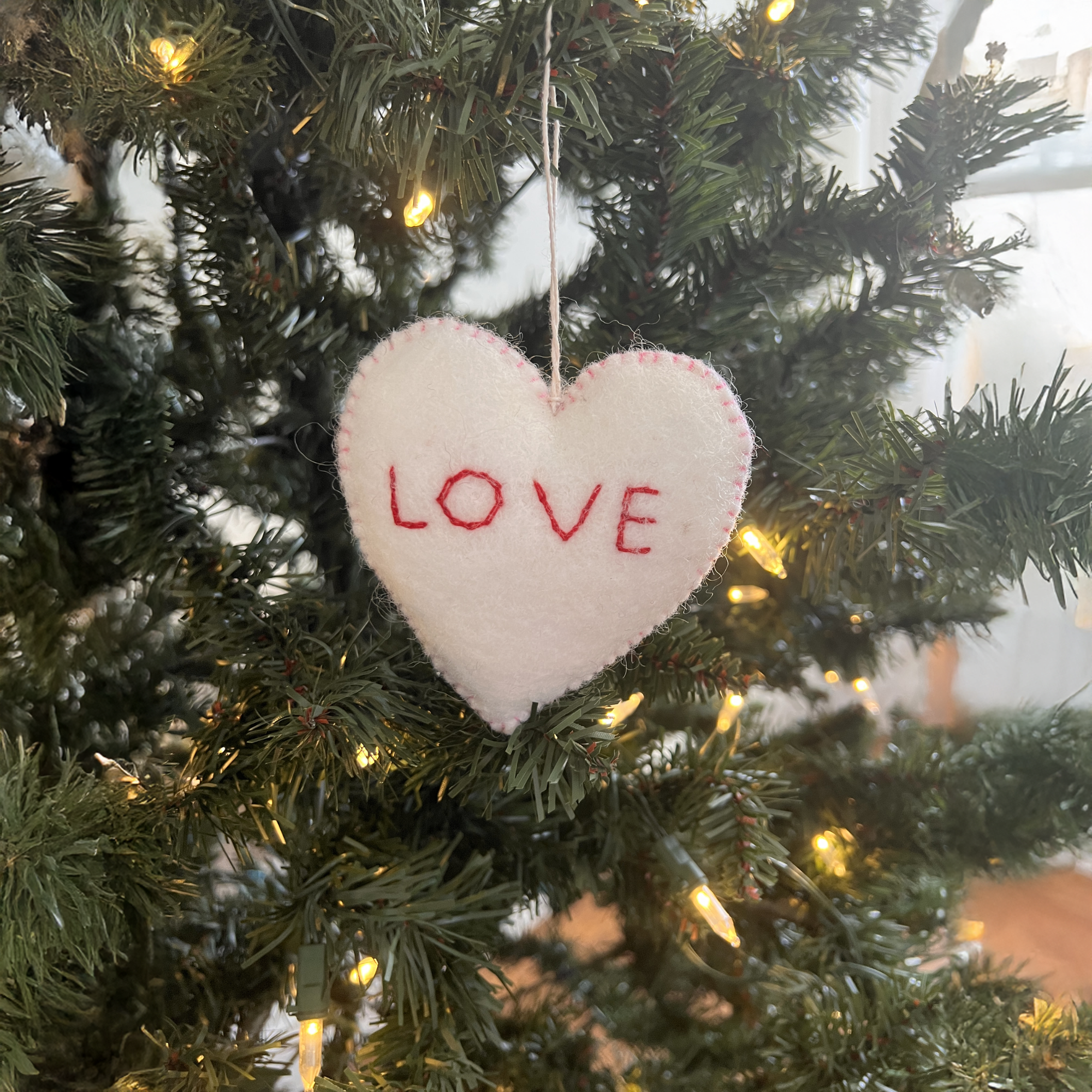 A trio of hand-stitched felt star and heart ornaments, crafted from sustainable wool, hangs on a Christmas tree adorned with white lights.