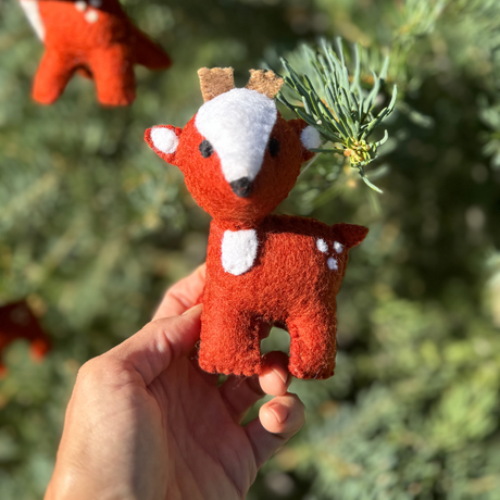 A hand holds one of the Trio of Hand-Stitched Felted Large Deer Ornaments, made from wool and perfect for decorating your tree, in front of green pine branches.
