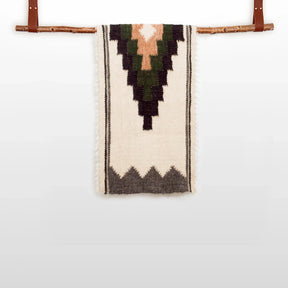 The Myr (Peace) Wool Bed / Couch Runner - Style A features handwoven geometric patterns and hangs from a wooden rod with leather straps, making it an ideal addition to sustainable home decor.