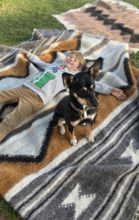 A smiling child lies on a patterned blanket outdoors, with a small black dog sitting nearby.
