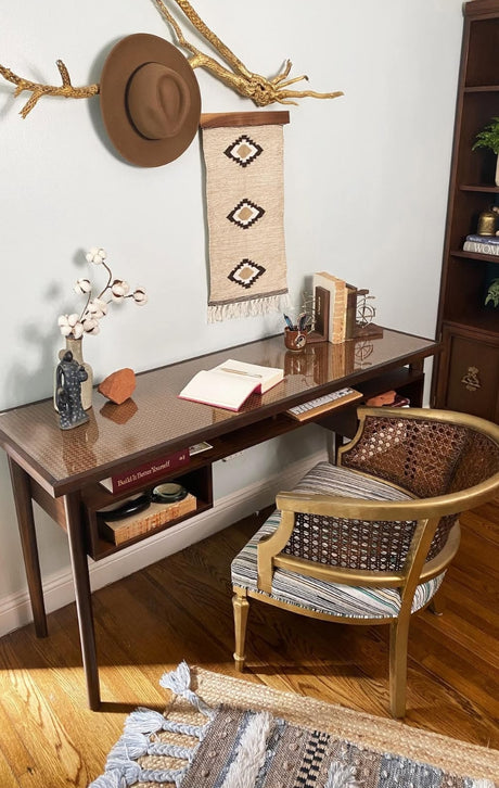 A cozy home office with a wooden desk, woven chair, books, and decorative items on a hardwood floor.