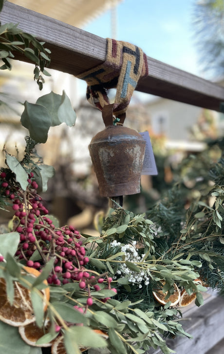 Rustic metal bell hanging on a wooden beam, surrounded by dried greenery, berries, and orange slices.