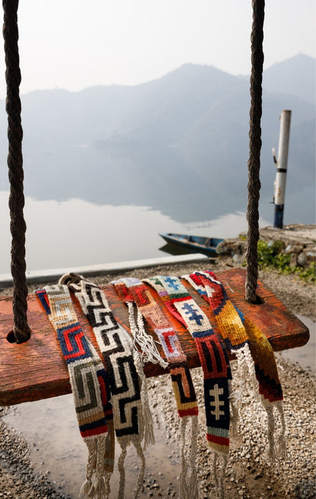 Colorful woven bracelets hang on a wooden swing by a calm lake with mountains in the background.