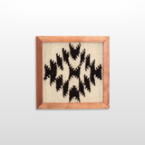 The Vira (Faith) Wool Framed Wall Art, made from undyed sheep’s wool, features a black geometric pattern on beige and is displayed against a plain white wall.