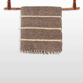 The Vivtsi (Sheep) Wool Statement Throw / Baby Blanket features a brown textured design with white horizontal stripes, hanging on a wooden rod with leather straps—ideal as a stylish accent for cozy spaces.