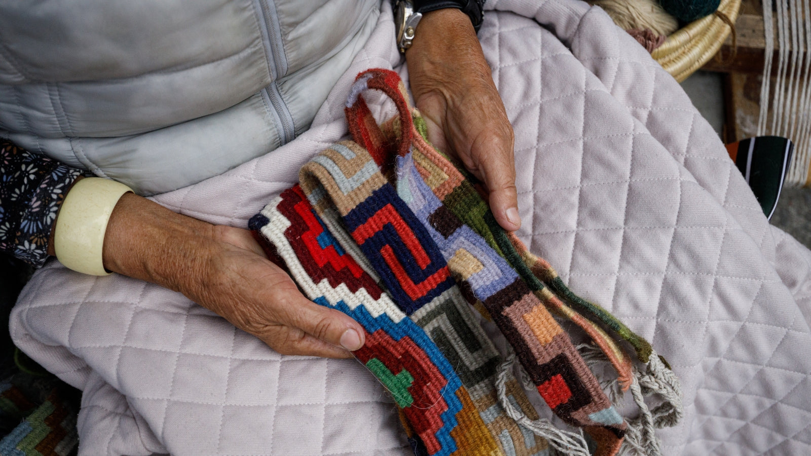 Elderly hands holding a colorful, geometric-patterned woven textile over a quilted blanket.