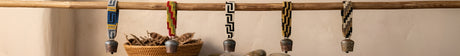 Five beaded necklaces with metal pendants hang from a wooden rod, above a woven basket with dried plants.