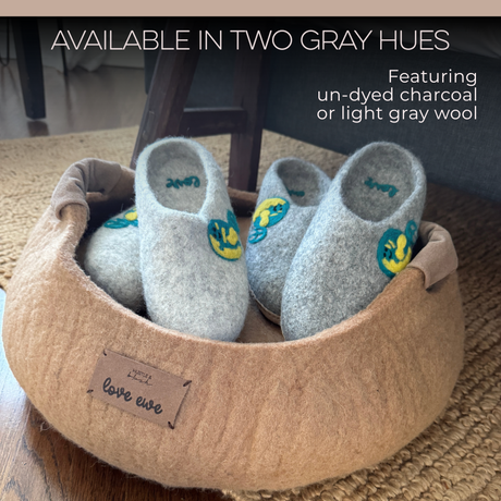 Love Ewe Hand-Felted Organic Wool Slippers for Men (Peace-LG), in gray with blue and yellow patches, are arranged in a soft brown basket beneath a chair.