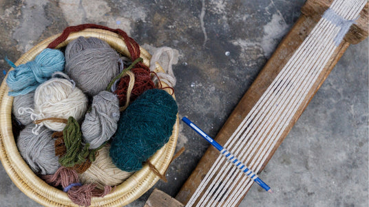 A basket of colorful yarn balls and a wooden loom with threads on a stone floor.