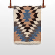 The Zirka (Star) Wool Statement Throw / Baby Blanket, handwoven with blue, brown, black, and cream geometric patterns, hangs on a wooden rod with leather straps.
