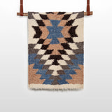The Zirka (Star) Wool Statement Throw / Baby Blanket, handwoven with blue, brown, black, and cream geometric patterns, hangs on a wooden rod with leather straps.