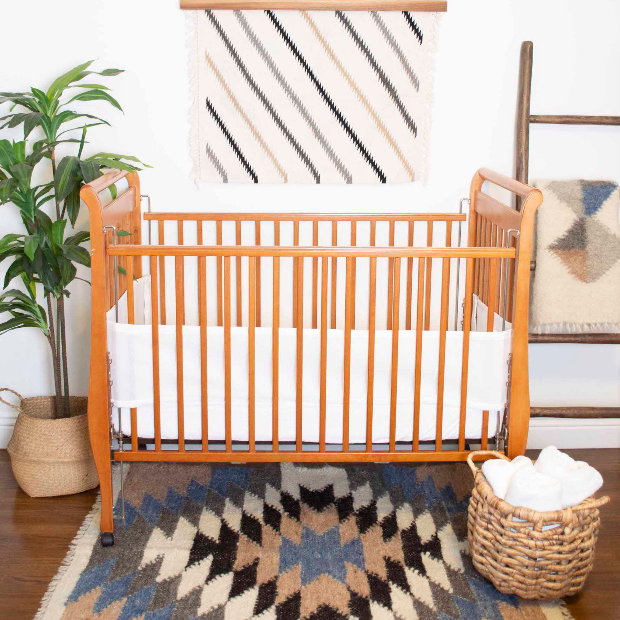 A wooden crib with white bedding sits on a patterned rug, alongside a plant and a basket containing the Zirka (Star) Wool Statement Throw / Baby Blanket.