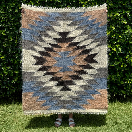 Person holding the Zirka (Star) Wool Statement Throw / Baby Blanket, featuring a geometric pattern and earthy tones, in front of lush green bushes and grass.