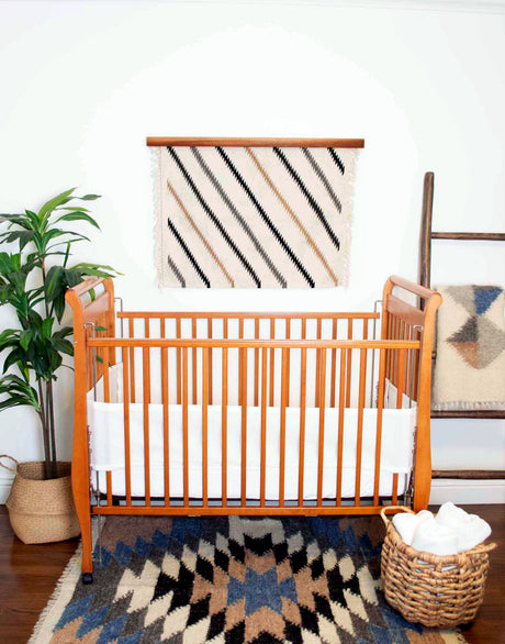 Cozy nursery with a wooden crib, white bedding, plant, blanket ladder, and eco-friendly decor featuring a Handmade in Ukraine Organic 100% Sheep Wool Woven Rug Wall Hanging.