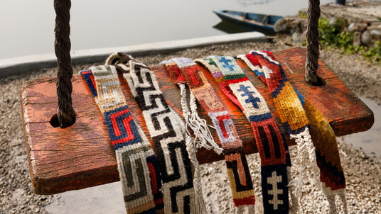 Colorful woven belts draped over a rustic wooden swing near a body of water with a boat in the background.