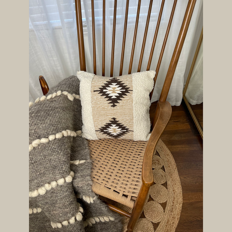 Wooden rocking chair with a woven seat, featuring the Handwoven Chitki (Crisp) Pillow and a chunky knit blanket draped over the arm.